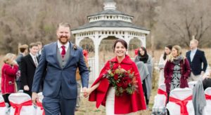 Happy grooom and bride in a white gown with a red cloak walking down aisle.